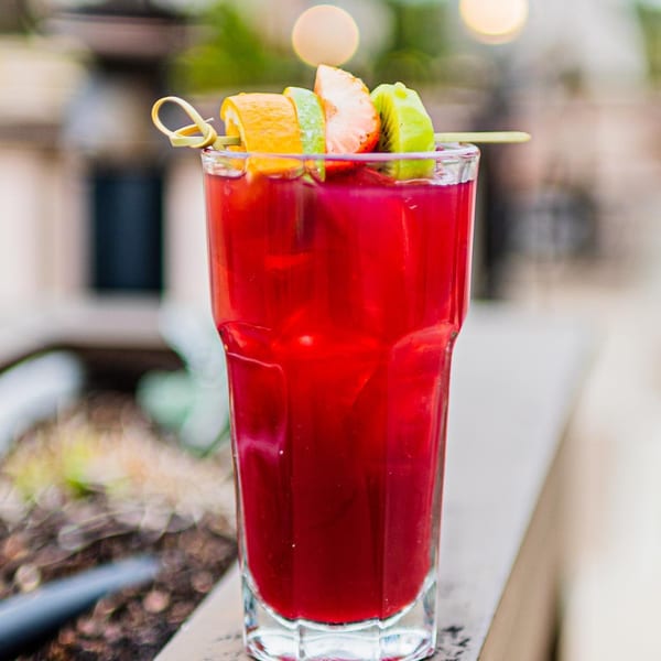 A vibrant red ZOCALO cocktail garnished with orange, strawberry, and kiwi slices on a skewer.