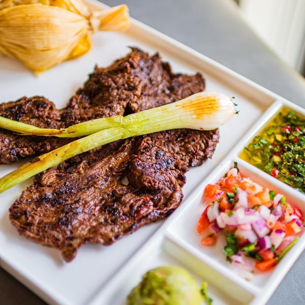 Grilled steak with green onions, tamale, pico de gallo, guacamole, and green sauce on a white plate.