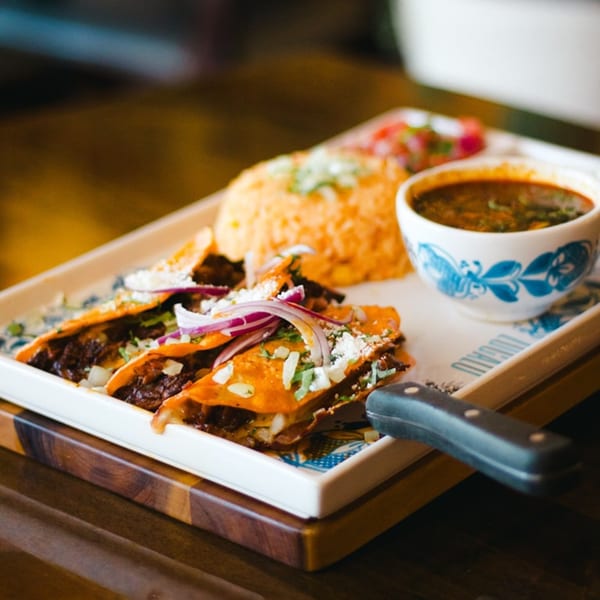 ZOCALO's birria tacos with rice, salsa, and a side of consommé on a white rectangular plate.