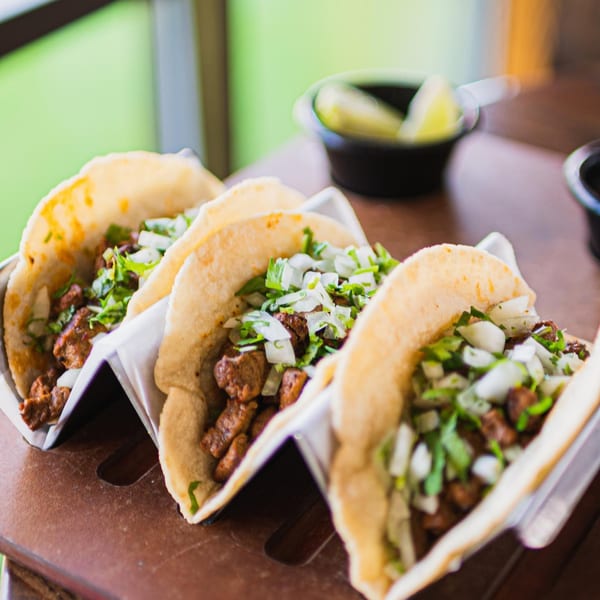 Three ZOCALO Mexican street tacos with meat, onions, and cilantro, with limes in the background.