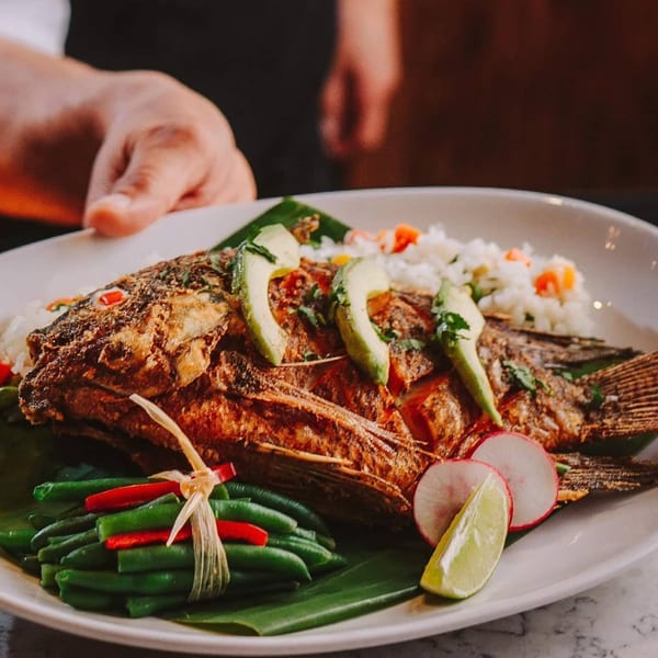 Whole fried fish with avocado, rice, green beans, radishes, and lime on a white plate.