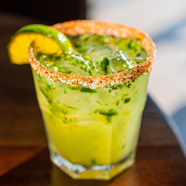 A ZOCALO Mexican margarita, light green with a chili-salt rim and lime wedge, on a dark table.